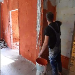 REMONTOLOGIA JACEK KRUPKA - Mężczyzna w kombinezonie maluje ceglaną ścianę na pomarańczowo w surowym wnętrzu, widoczne przewody elektryczne i wiadro z farbą.