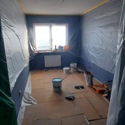 REMONTOLOGIA JACEK KRUPKA - Przygotowanie pokoju do malowania: ściany zabezpieczone folią, podłoga wyłożona kartonem, widoczne pędzle, wałki i wiaderka z farbą.