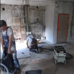 REMONTOLOGIA JACEK KRUPKA - Dwóch mężczyzn w kombinezonach roboczych podczas remontu pokoju: jeden zbiera gruz do worka, drugi szlifuje podłogę przy użyciu szlifierki podłączonej do odkurzacza przemysłowego; widoczne surowe...