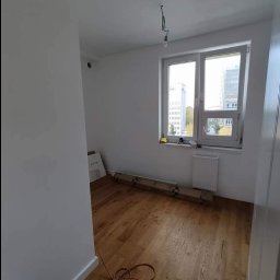 REMONTOLOGIA JACEK KRUPKA - Pokój po remoncie z nową drewnianą podłogą, białe ściany, okno z widokiem na budynki, zwisający kabel od lampy, kartony z panelami pod ścianą.