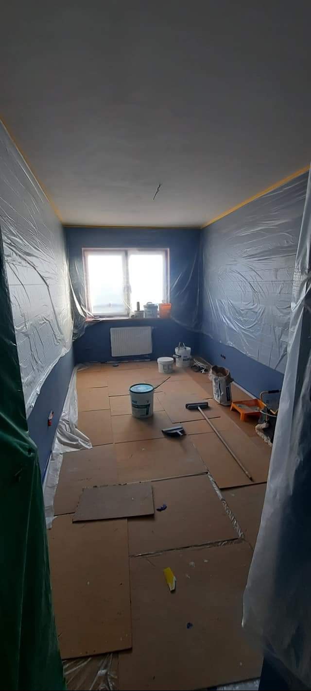 Przygotowanie pokoju do malowania: ściany zabezpieczone folią, podłoga wyłożona kartonem, widoczne pędzle, wałki i wiaderka z farbą.