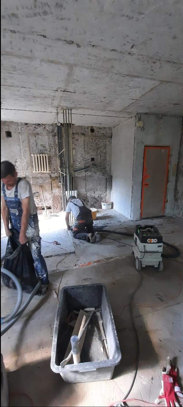 Dwóch mężczyzn w kombinezonach roboczych podczas remontu pokoju: jeden zbiera gruz do worka, drugi szlifuje podłogę przy użyciu szlifierki podłączonej do odkurzacza przemysłowego; widoczne surowe...