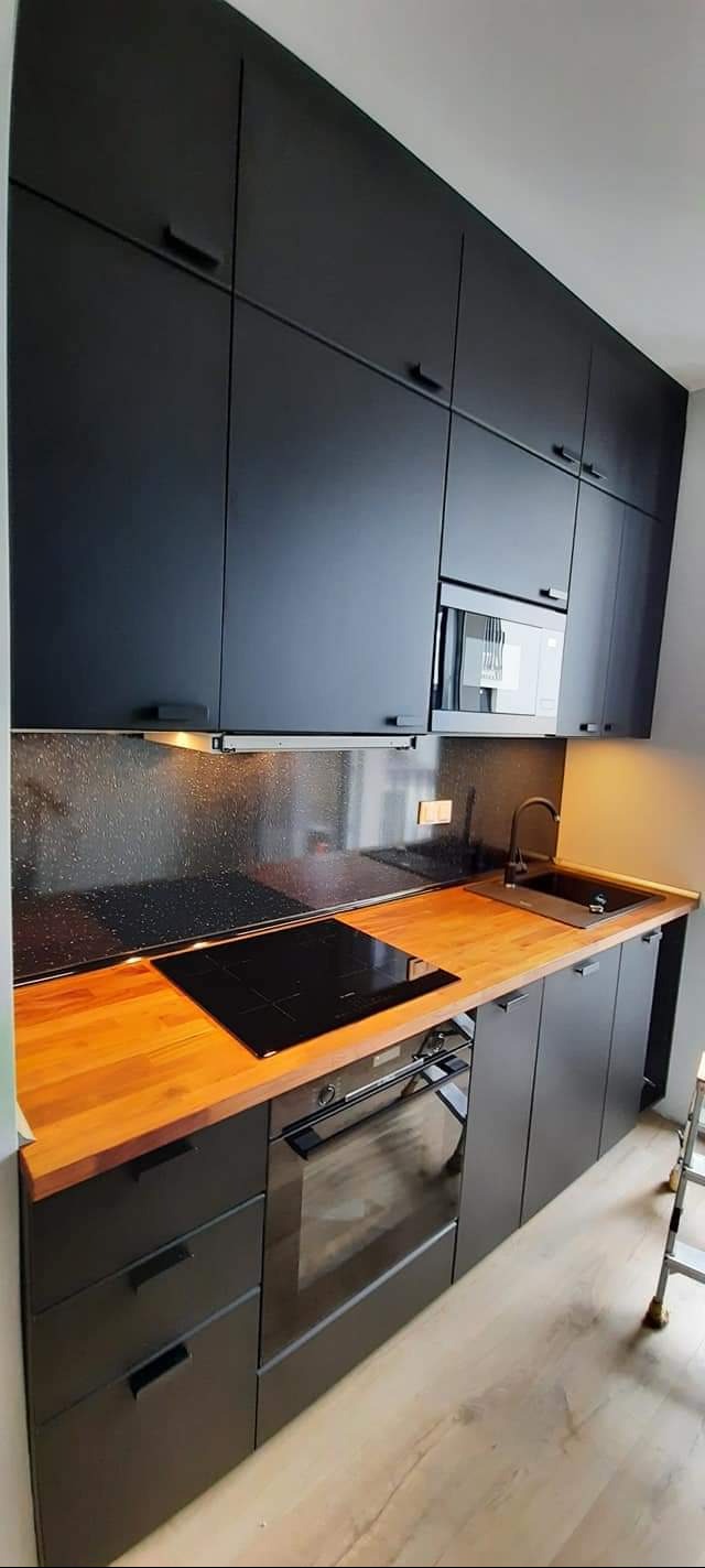 Czarna, nowoczesna kuchnia z drewnianym blatem, płytą indukcyjną, piekarnikiem i mikrofalówką w zabudowie, oświetlona punktowo pod szafkami.