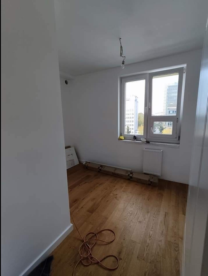 Pokój po remoncie z nową drewnianą podłogą, białe ściany, okno z widokiem na budynki, zwisający kabel od lampy, kartony z panelami pod ścianą.