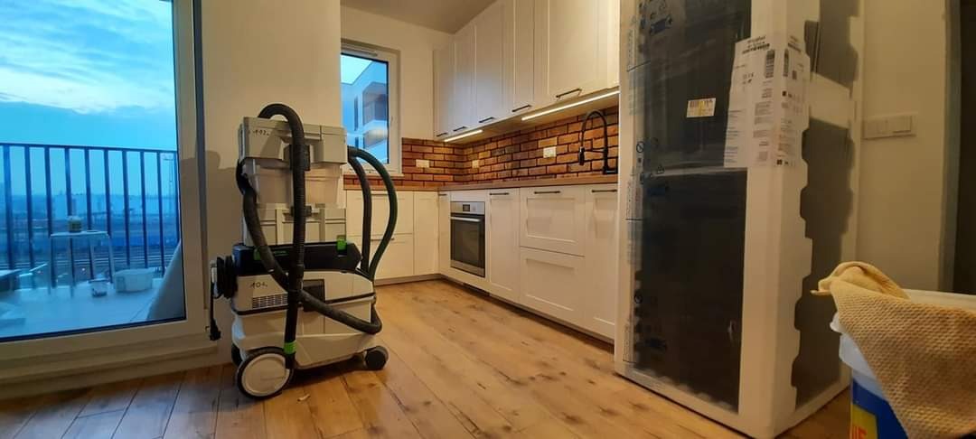 Wnętrze kuchni w trakcie prac wykończeniowych, widoczny odkurzacz przemysłowy Festool, białe szafki kuchenne z ceglaną ścianą nad blatem, drewniana podłoga i częściowo zakryta lodówka.