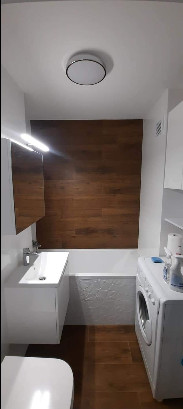 Nowoczesna łazienka z brązowymi panelami ściennymi imitującymi drewno, białą umywalką z szafką, podświetlanym lustrem, pralką i płytkami podłogowymi w tym samym odcieniu brązu.