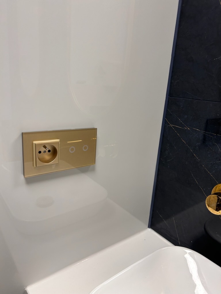 Biała ściana z połyskiem, złote gniazdko elektryczne i dotykowy włącznik światła, fragment umywalki i czarny marmur na ścianie w tle.
