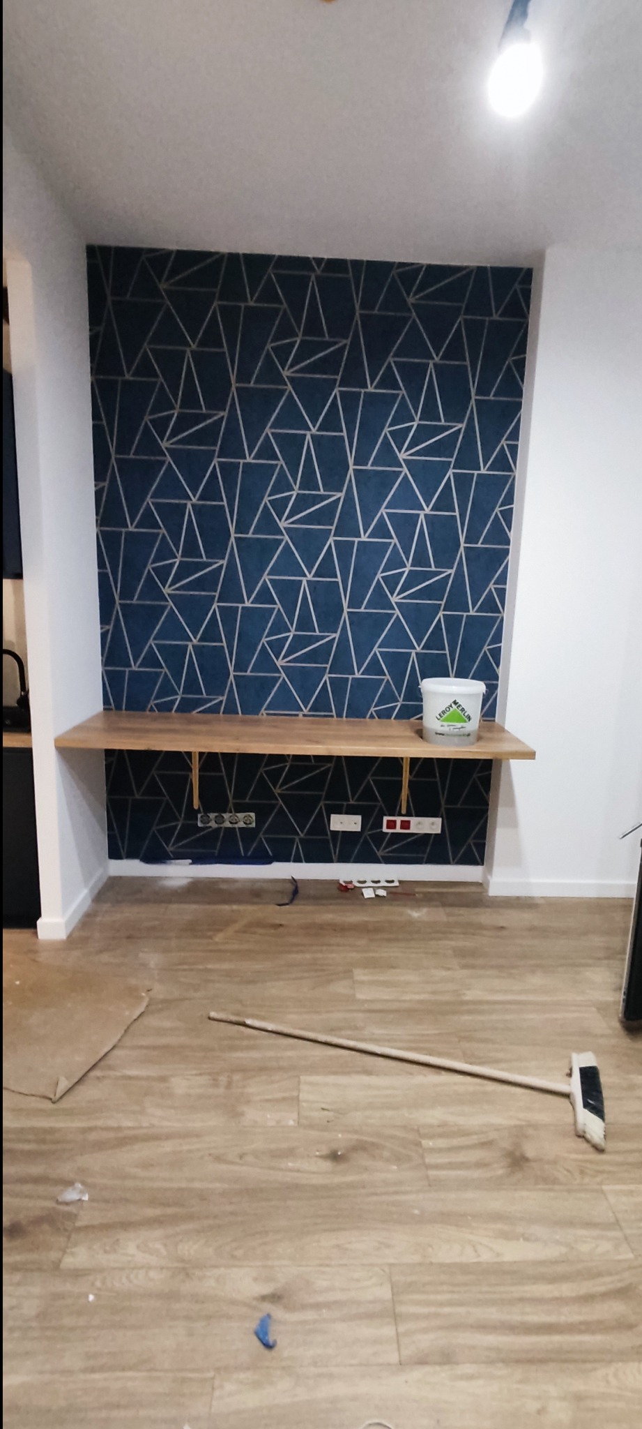Ściana z tapetą w geometryczne wzory, drewniana półka wsparta na wspornikach, widoczne gniazdka elektryczne, podłoga z panelami, białe wiadro z logo sklepu budowlanego, pomieszczenie w trakcie prac...