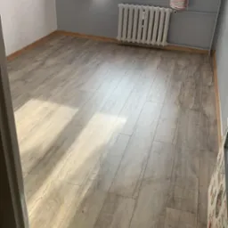 Jasne panele podłogowe imitujące drewno, ułożone w pokoju z szarą ścianą i oknem z pomarańczowo-białymi zasłonami, widoczny fragment grzejnika i pr&oacute;g drzwiowy.