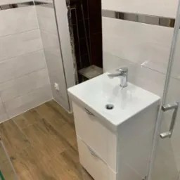 Nowoczesna biała szafka umywalkowa z chromowaną baterią w łazience z beżowymi płytkami imitującymi drewno na podłodze i białymi płytkami ściennymi z dekoracyjnym paskiem