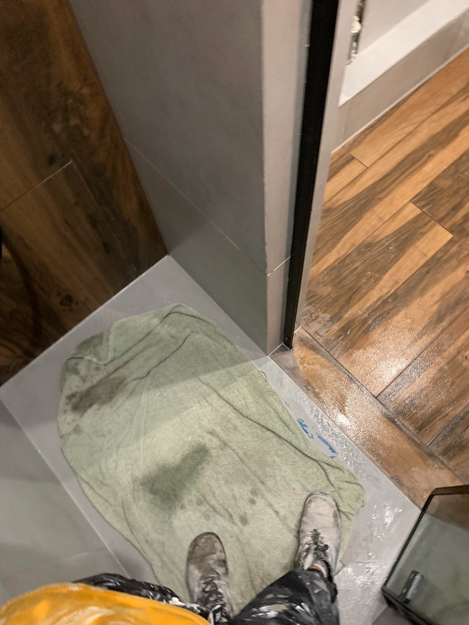 Podłoga łazienki po fugowaniu, mokry ręcznik i buty robocze na świeżo położonych szarych płytkach, widoczne drewnopodobne płytki na ścianie i podłodze kabiny prysznicowej.