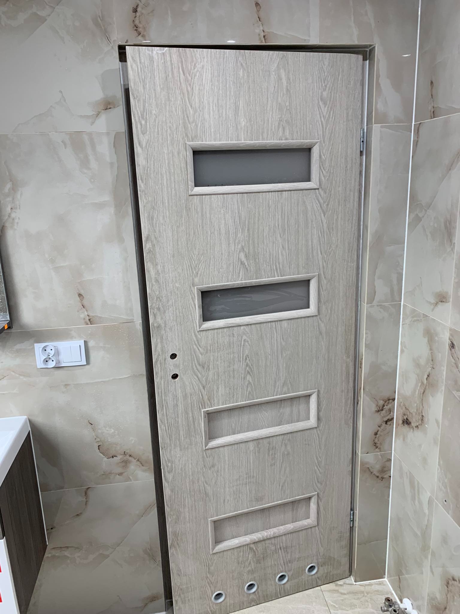 Jasna, drewnopodobna drzwi wewnętrzne z czterema prostokątnymi wstawkami, z których dwie górne są przeszklone, częściowo zamontowane w łazience z marmurkowymi płytkami.