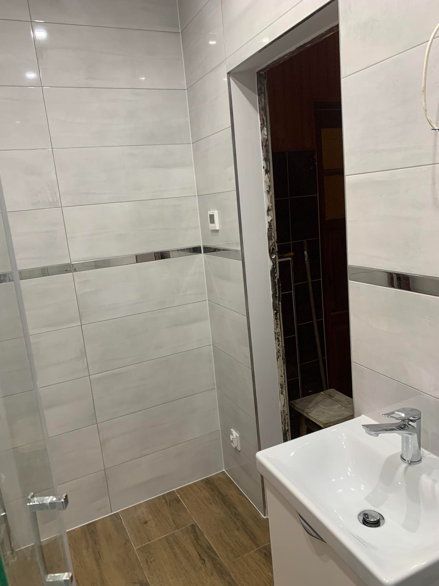 Częściowo wykończona łazienka z białymi płytkami ściennymi z metalicznymi paskami, drewnopodobną podłogą, białą umywalką z chromowaną baterią i widoczną futryną drzwi w trakcie montażu.