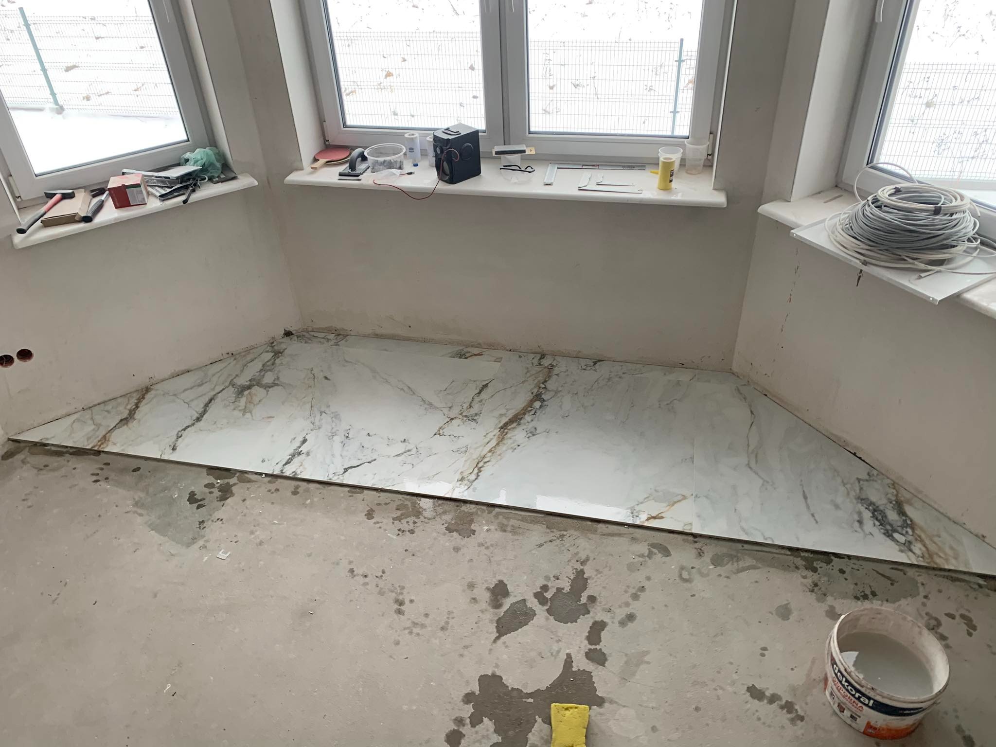 Fragment podłogi z ułożonymi płytkami imitującymi marmur, widoczne ślady po zaprawie, wiadro z farbą na posadzce, okna z widokiem na zewnątrz, narzędzia na parapecie.