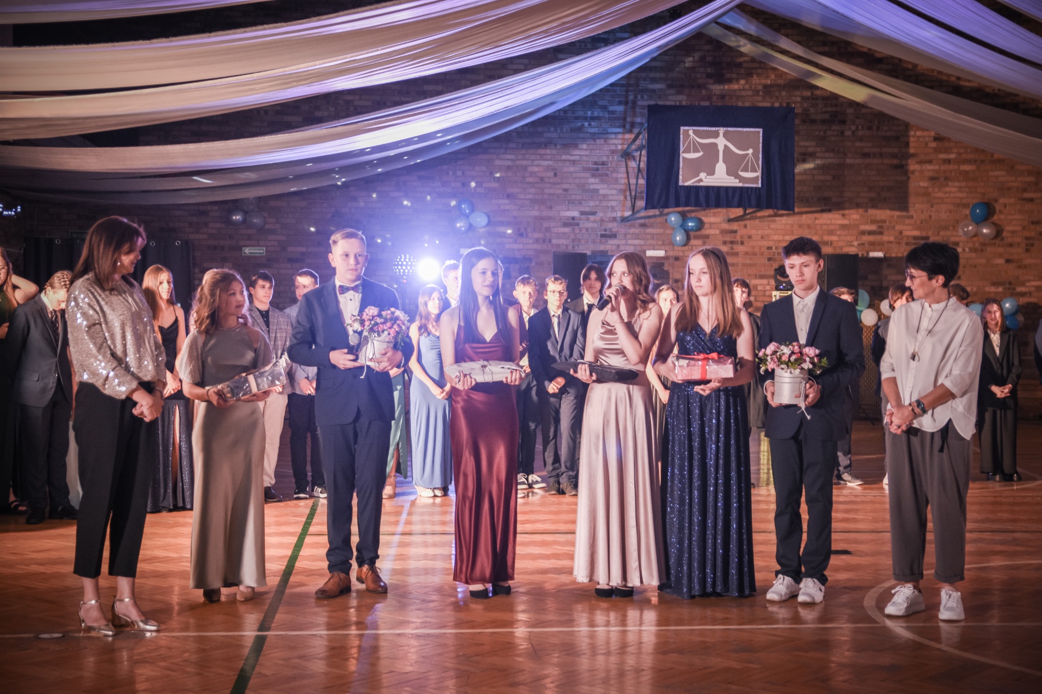 Uczniowie w strojach galowych, stojący na scenie podczas uroczystości szkolnej, trzymający upominki i kwiaty, w tle dekoracje z materiału i balonów.