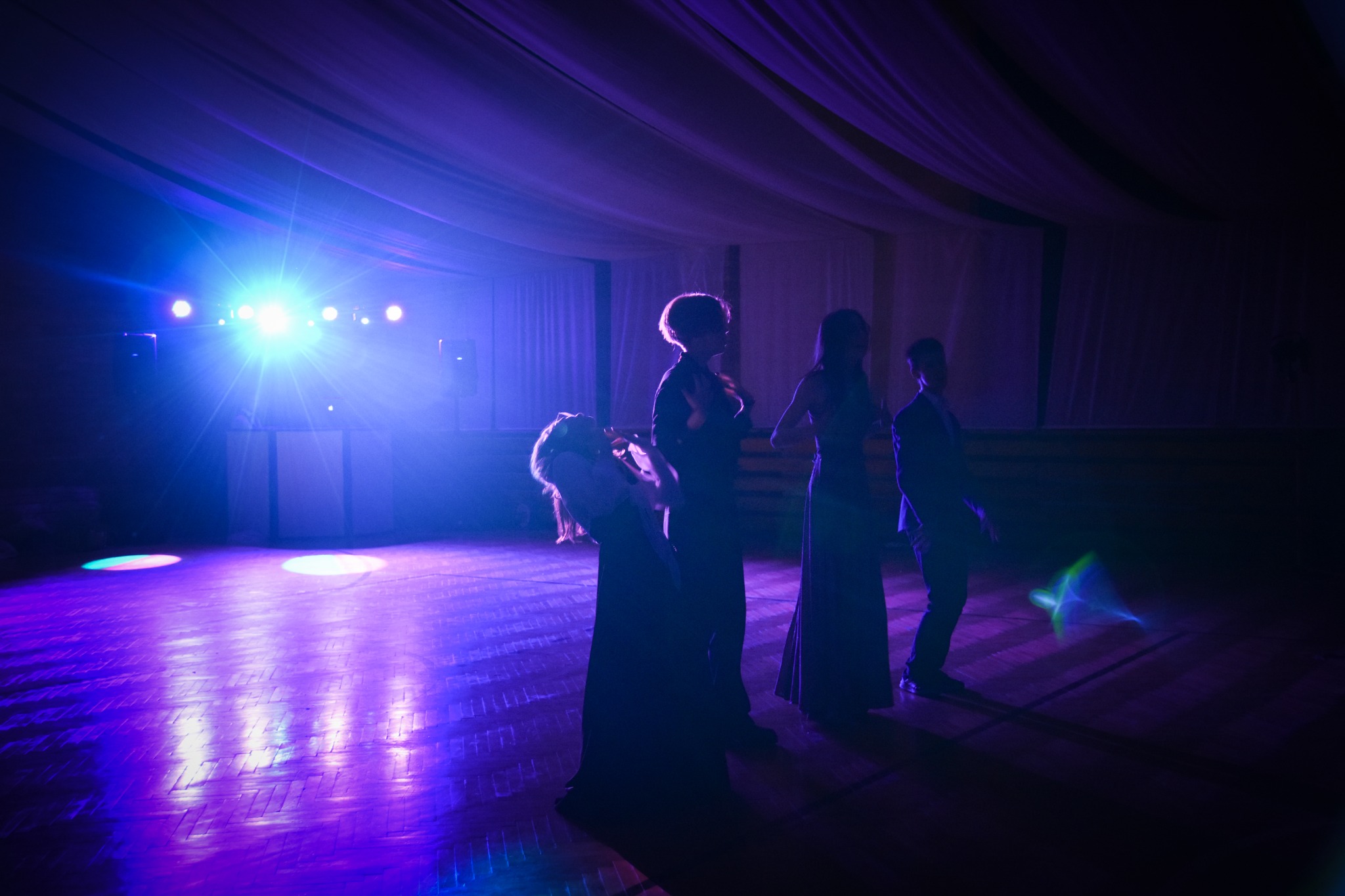 Silhouettes of four people on a dance floor illuminated by blue and purple stage lighting, with a DJ setup visible in the background.