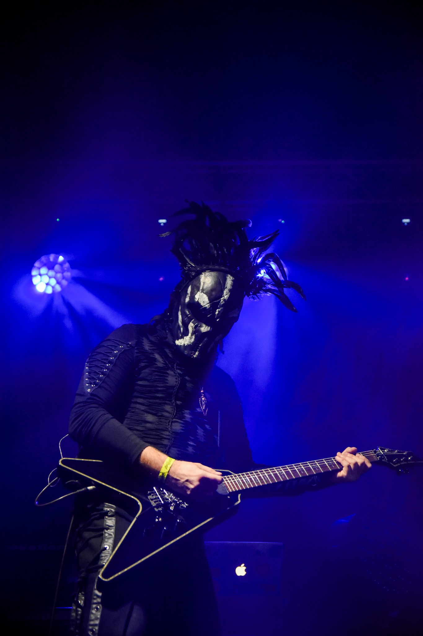 Postać w masce z upiornym makijażem grająca na gitarze elektrycznej podczas koncertu, oświetlona niebieskim światłem.