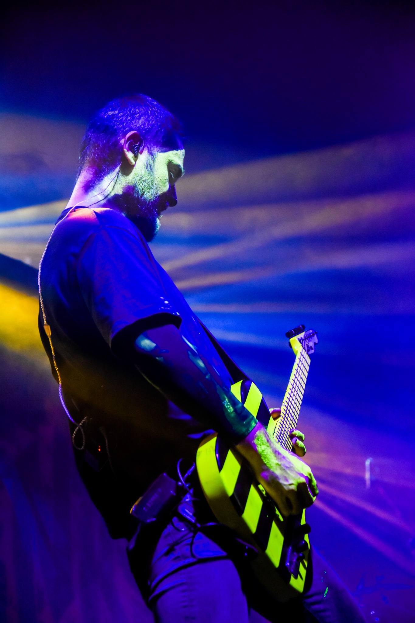 Gitarzysta z brodą gra na żółto-czarnej gitarze elektrycznej na scenie oświetlonej niebieskim i żółtym światłem.