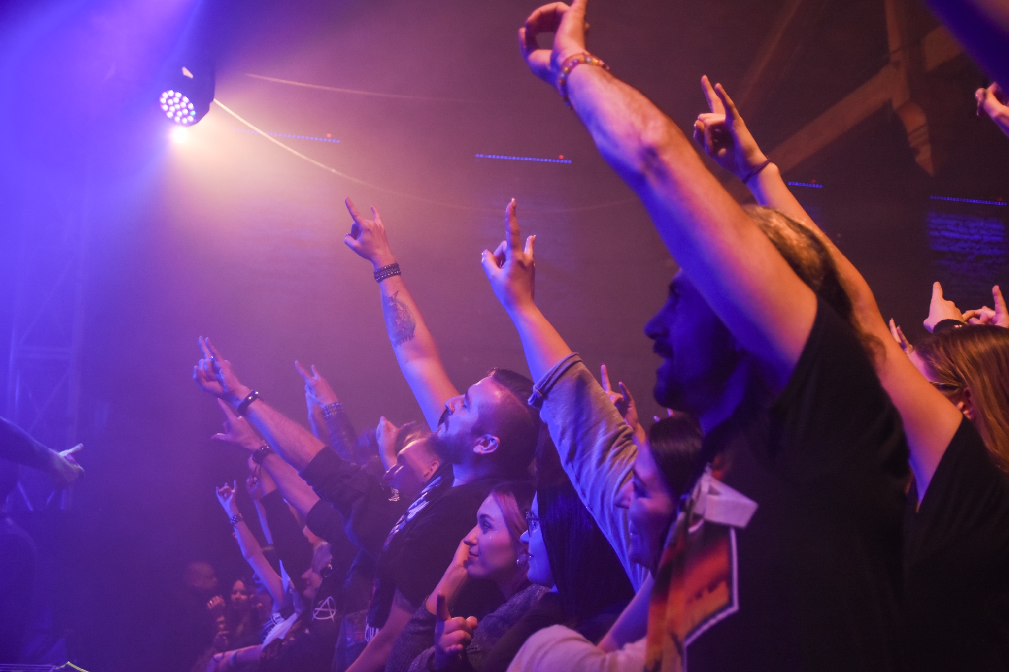 Rozświetlony tłum na koncercie, uniesione ręce w geście aprobaty, oświetlenie sceniczne w odcieniach fioletu i pomarańczy, widoczny fragment sceny i oświetlenia punktowego.