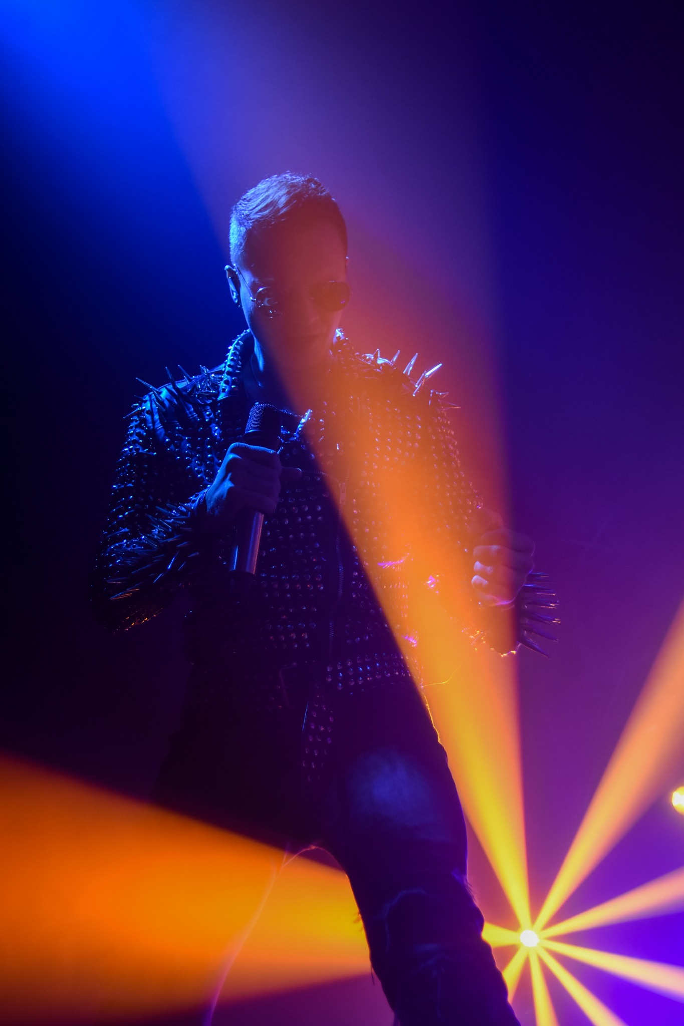 Sylwetka piosenkarza z mikrofonem w ręku, ubrana w skórzaną kurtkę z ćwiekami, oświetlona niebieskim i pomarańczowym światłem scenicznym.