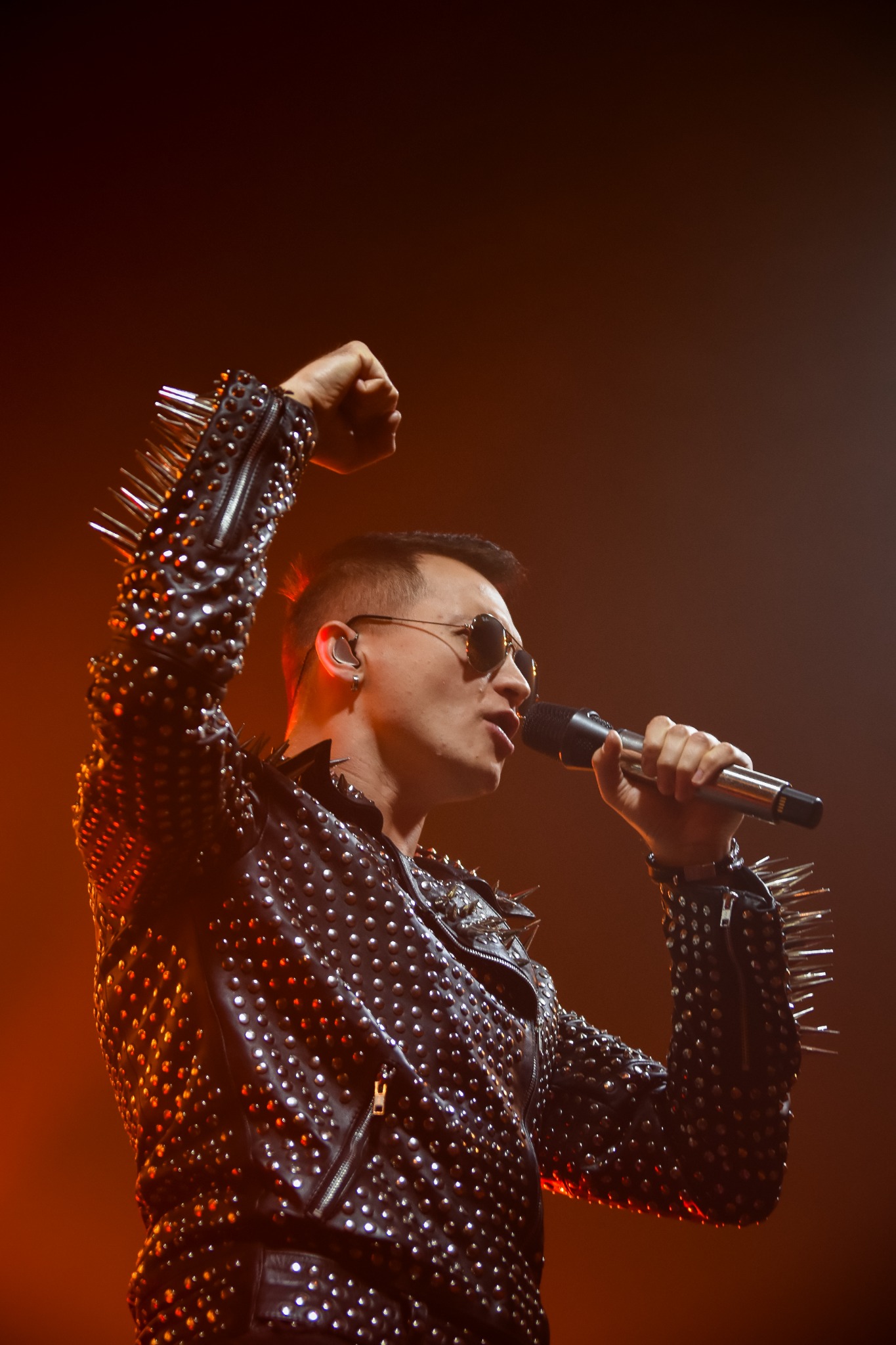 Wokalista w skórzanej kurtce z ćwiekami i okularach przeciwsłonecznych śpiewa do mikrofonu na scenie w pomarańczowym oświetleniu, unosząc pięść w geście triumfu.