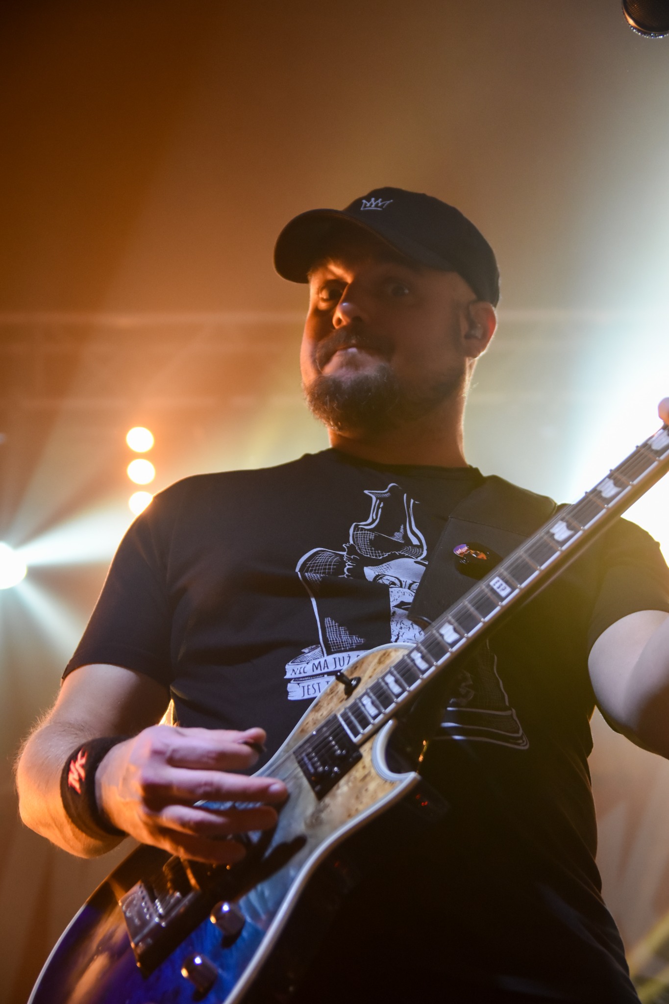 Gitarzysta w czarnej koszulce z nadrukiem i czapce z daszkiem gra na gitarze elektrycznej podczas koncertu, oświetlony pomarańczowym światłem.