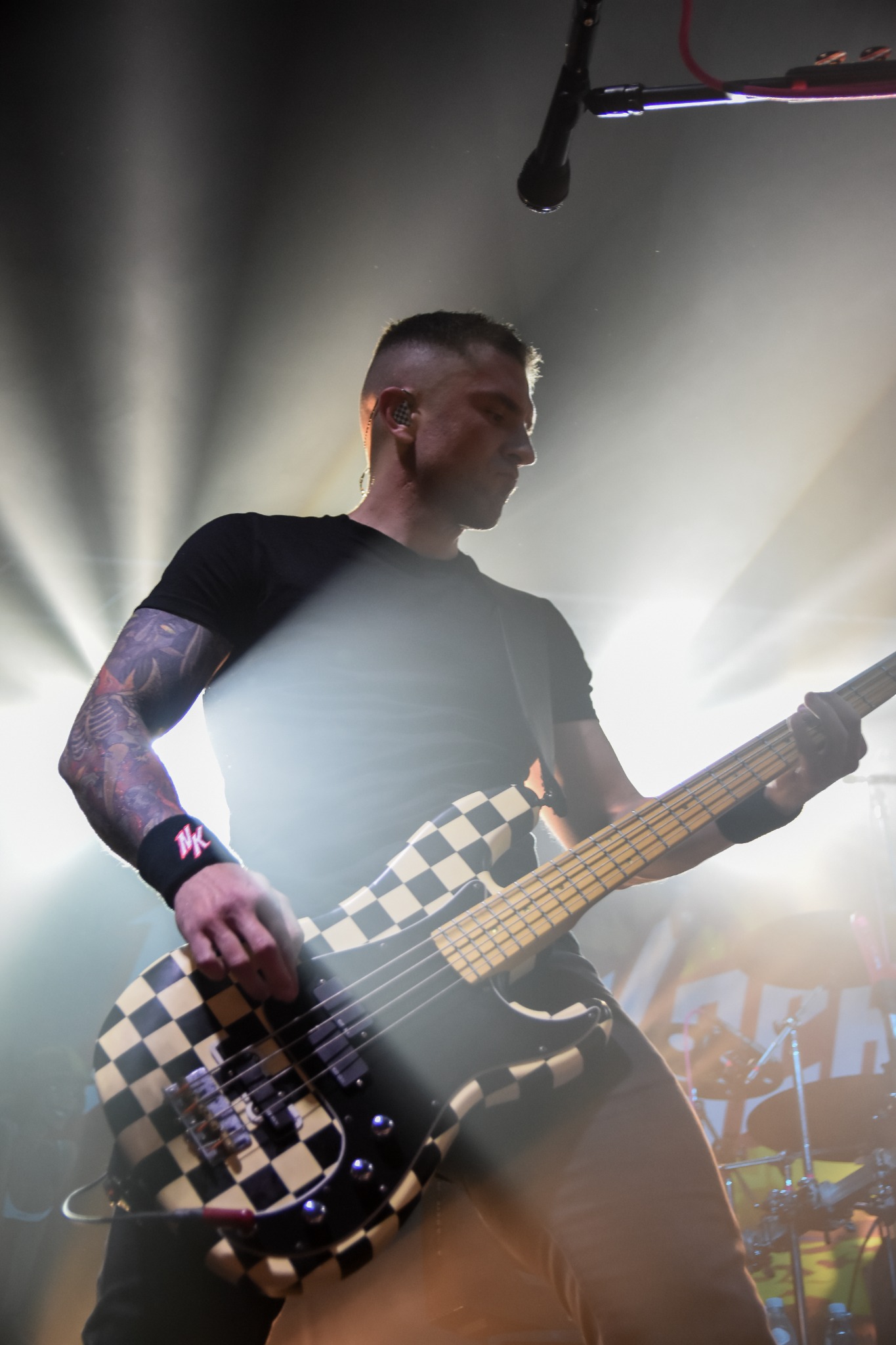 Basista z tatuażami na ramieniu gra na gitarze basowej w czarno-białą szachownicę podczas koncertu, oświetlony przez reflektory.