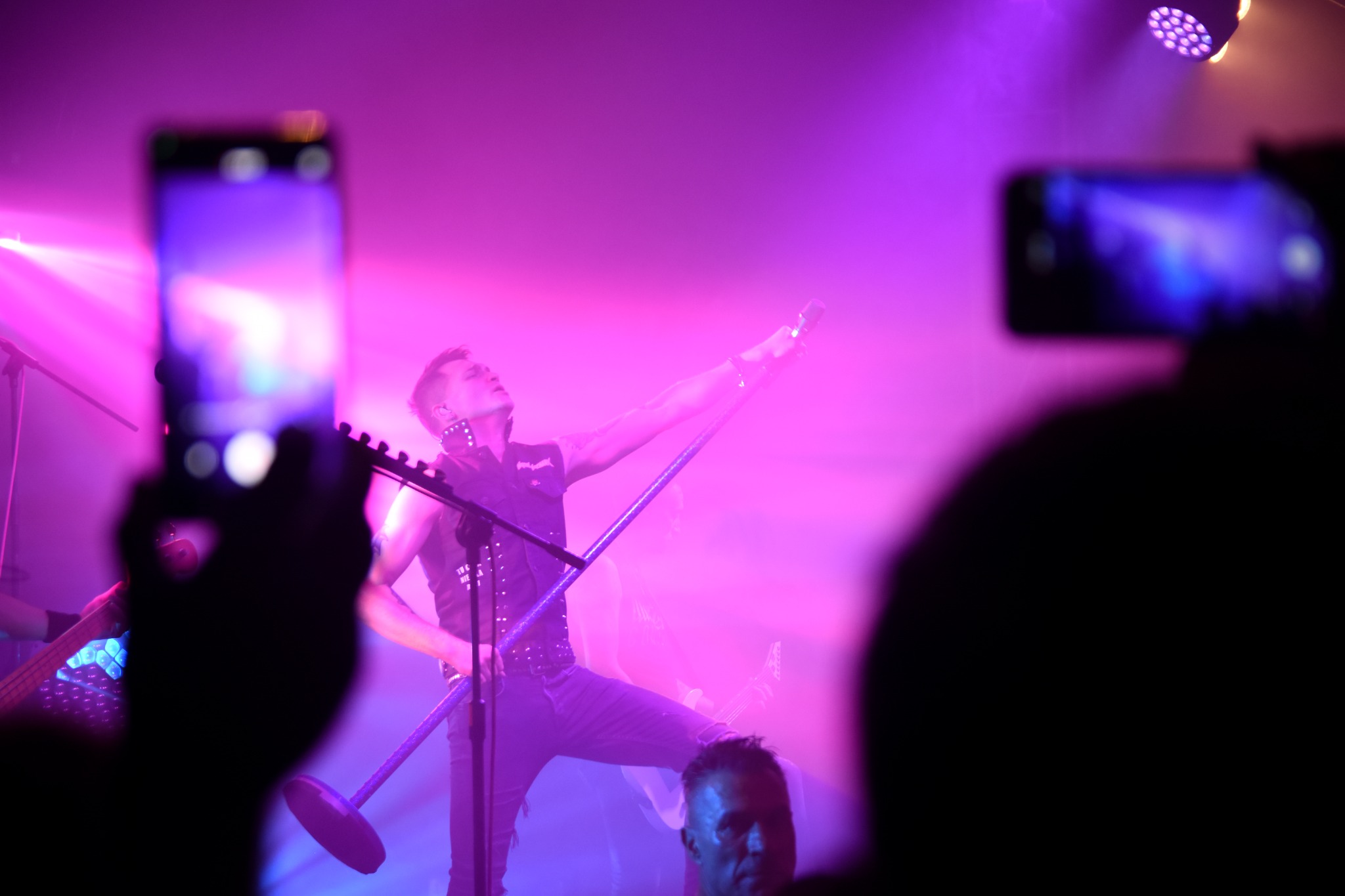 Wokalista z mikrofonem na statywie, w skórzanej kamizelce, oświetlony fioletowymi światłami sceny, widziany zza telefonów nagrywających publiczności.
