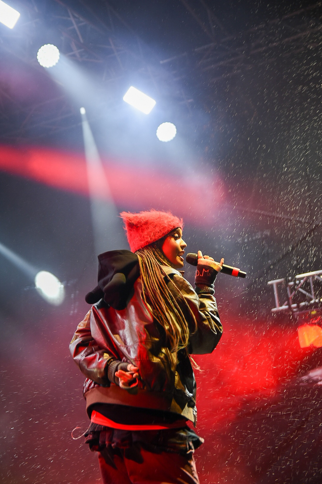 Piosenkarka z dredami w czerwonej czapce, w skórzanej kurtce, śpiewa do mikrofonu na scenie w strugach deszczu, oświetlona reflektorami.