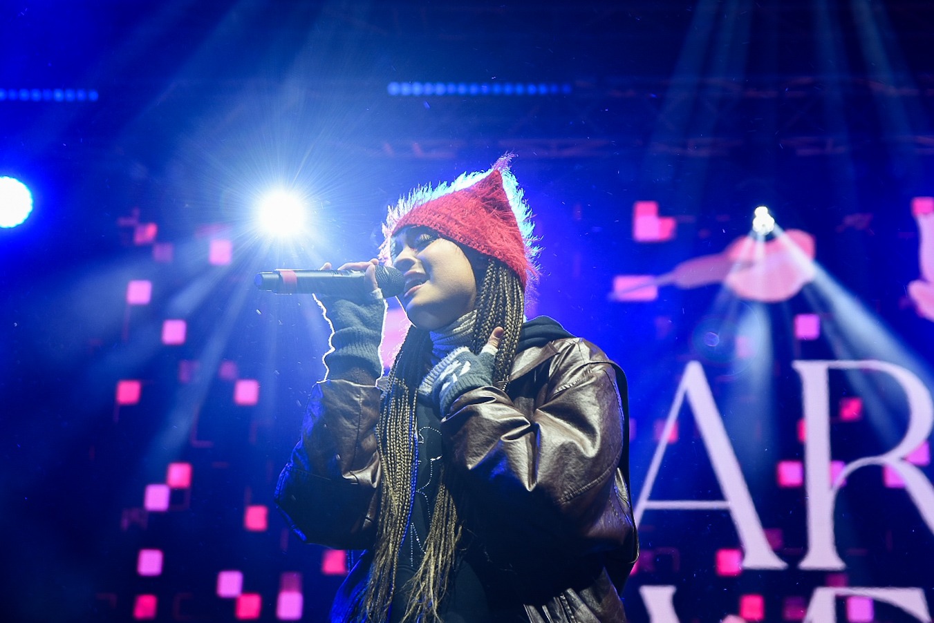 Młoda kobieta z warkoczykami w czerwonej czapce śpiewa do mikrofonu na scenie oświetlonej reflektorami.