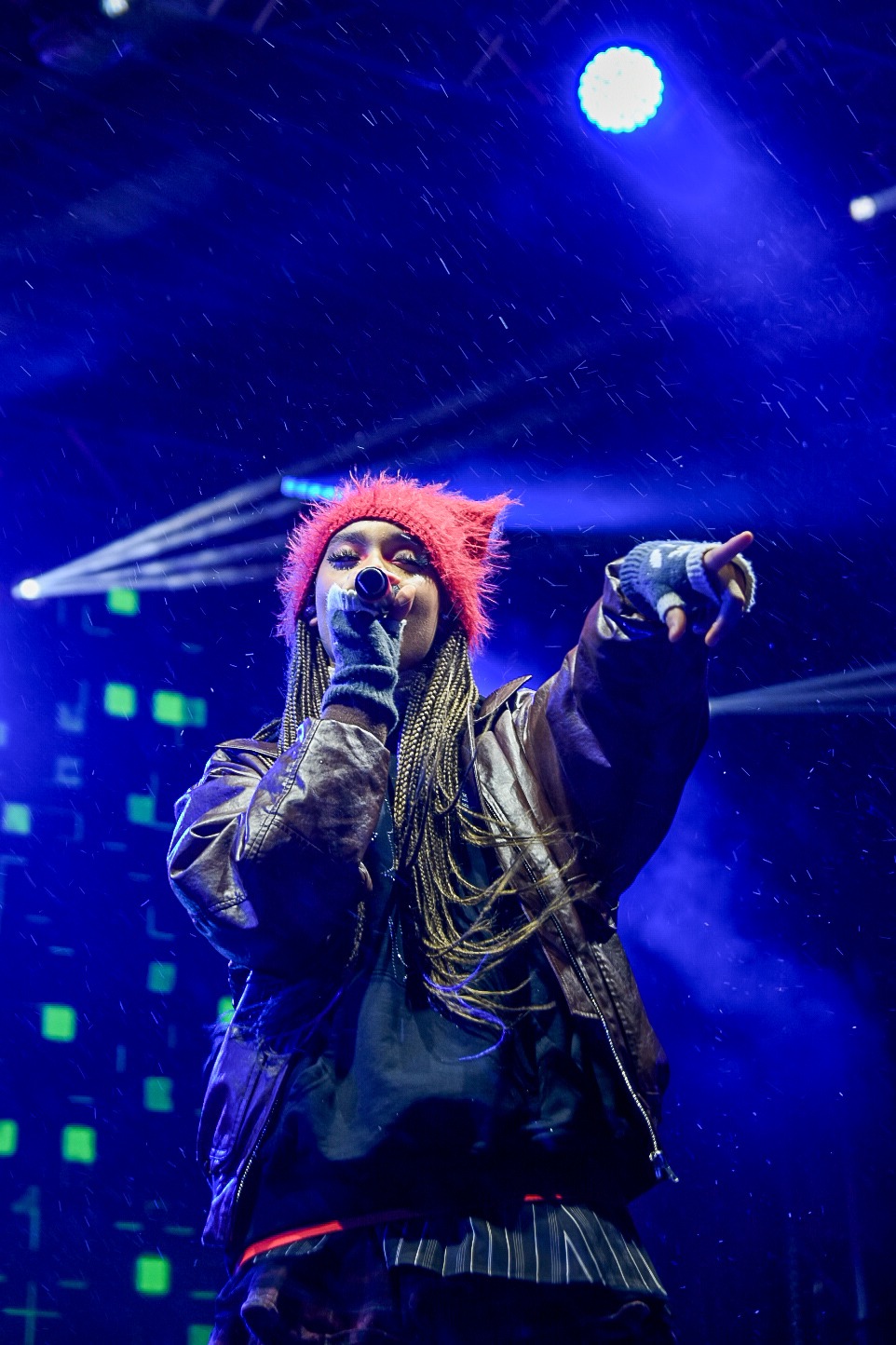 Piosenkarka z warkoczami w czerwonej czapce i skórzanej kurtce śpiewa do mikrofonu na scenie podczas wydarzenia oświetlonego niebieskim światłem.