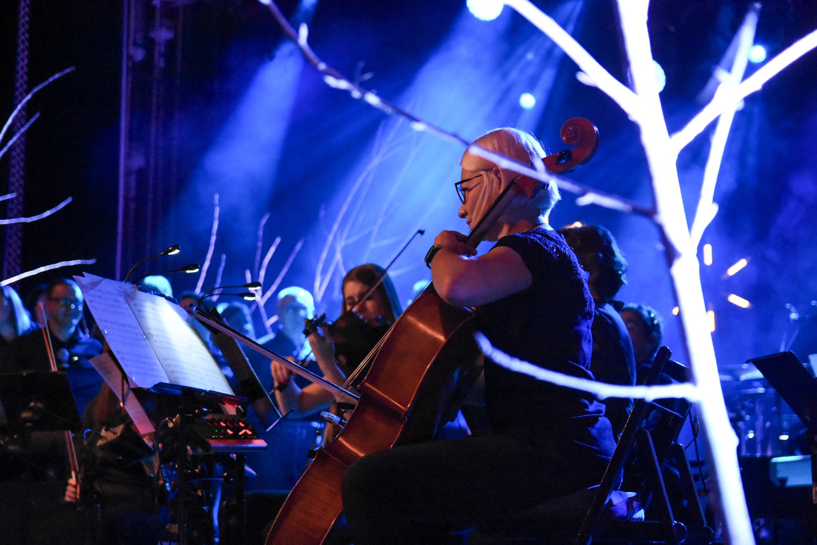 Orkiestra symfoniczna w niebieskim oświetleniu: wiolonczelistka w okularach gra na instrumencie, obok skrzypaczka i pulpity z nutami.