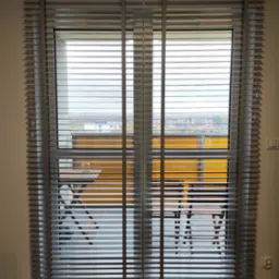 Szare poziome lamele okienne z widokiem na balkon z meblami i zabudową w tle.