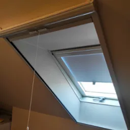 Szara roleta wewnętrzna zamontowana w oknie dachowym, częściowo zasłonięta, z widocznym mechanizmem sterującym sznurkiem.