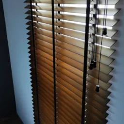 Zamknięte drewniane żaluzje z czarnymi sznurkami i obciążnikami, widoczne z boku, z podłogą z paneli w dolnej części kadru.