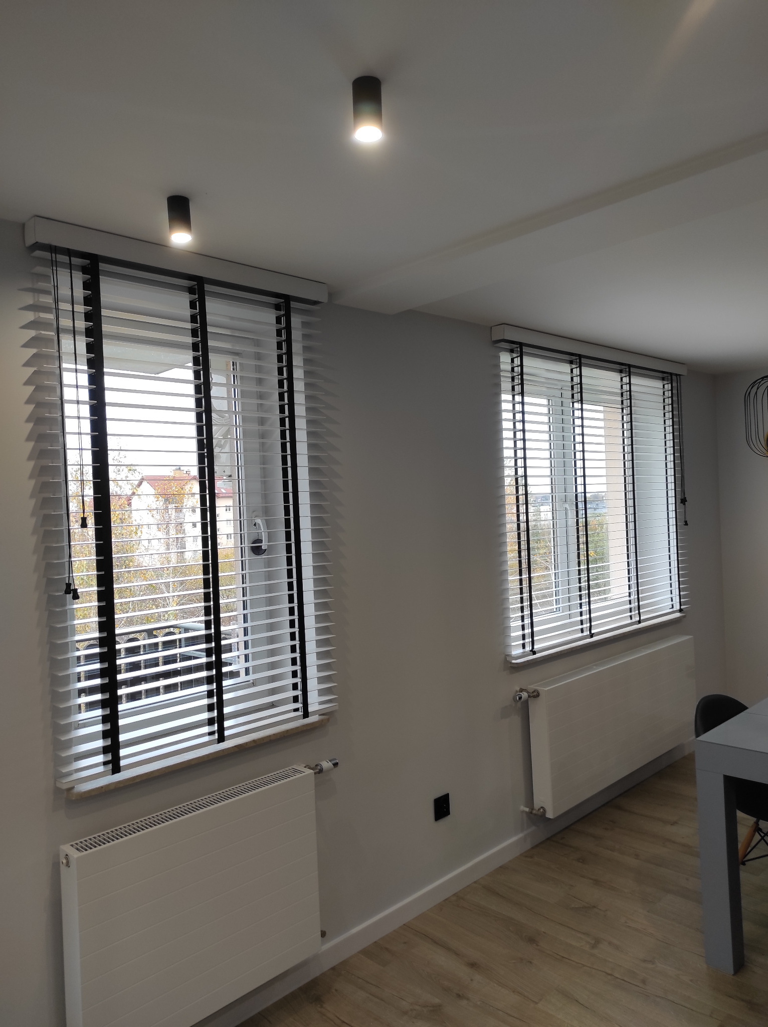 Dwa okna z białymi żaluzjami z czarnymi detalami w nowoczesnym wnętrzu, widok na budynki za oknem.