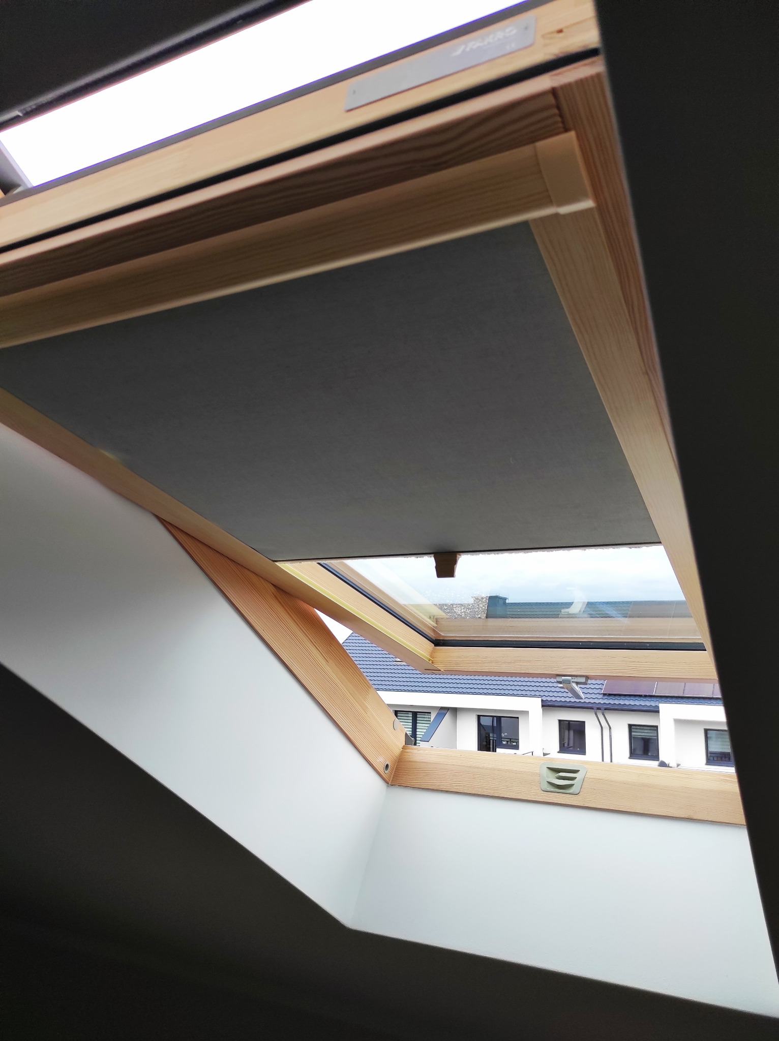 Szara tkanina zaciemniająca w oknie dachowym z drewnianą ramą, widok na sąsiednie domy z niebieskimi dachami.