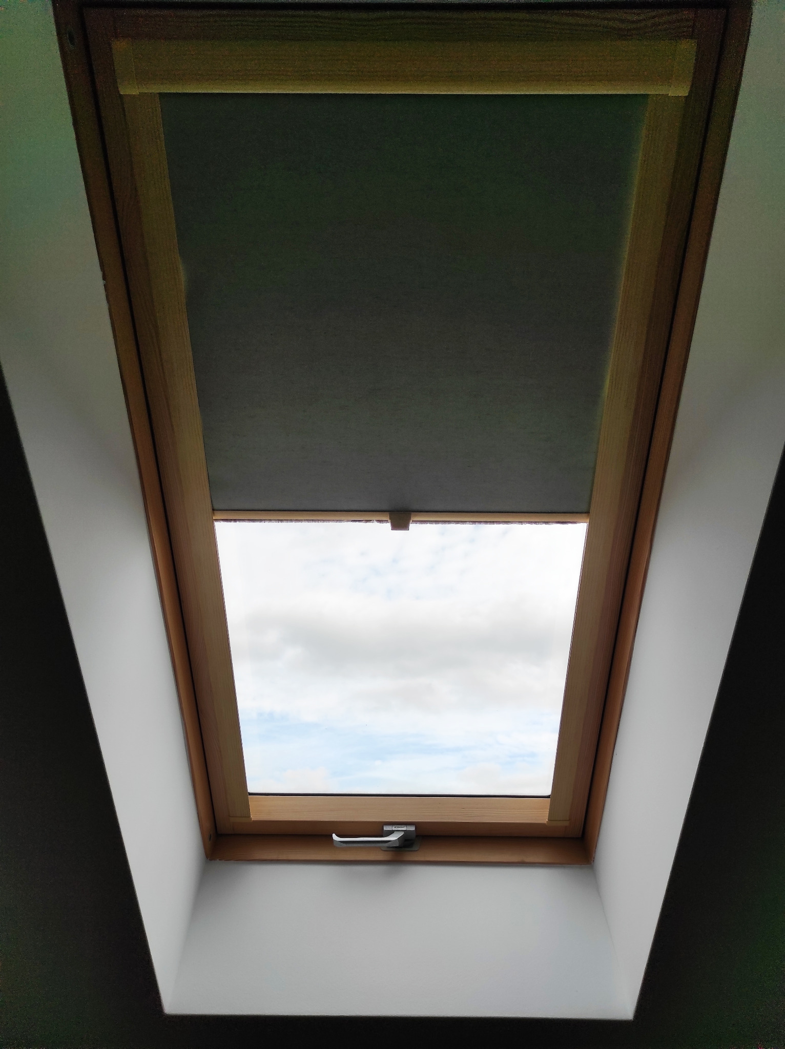 Okno dachowe z drewnianą ramą i częściowo zasuniętą, szarą roletą wpadającą w cień, widok na pochmurne niebo.