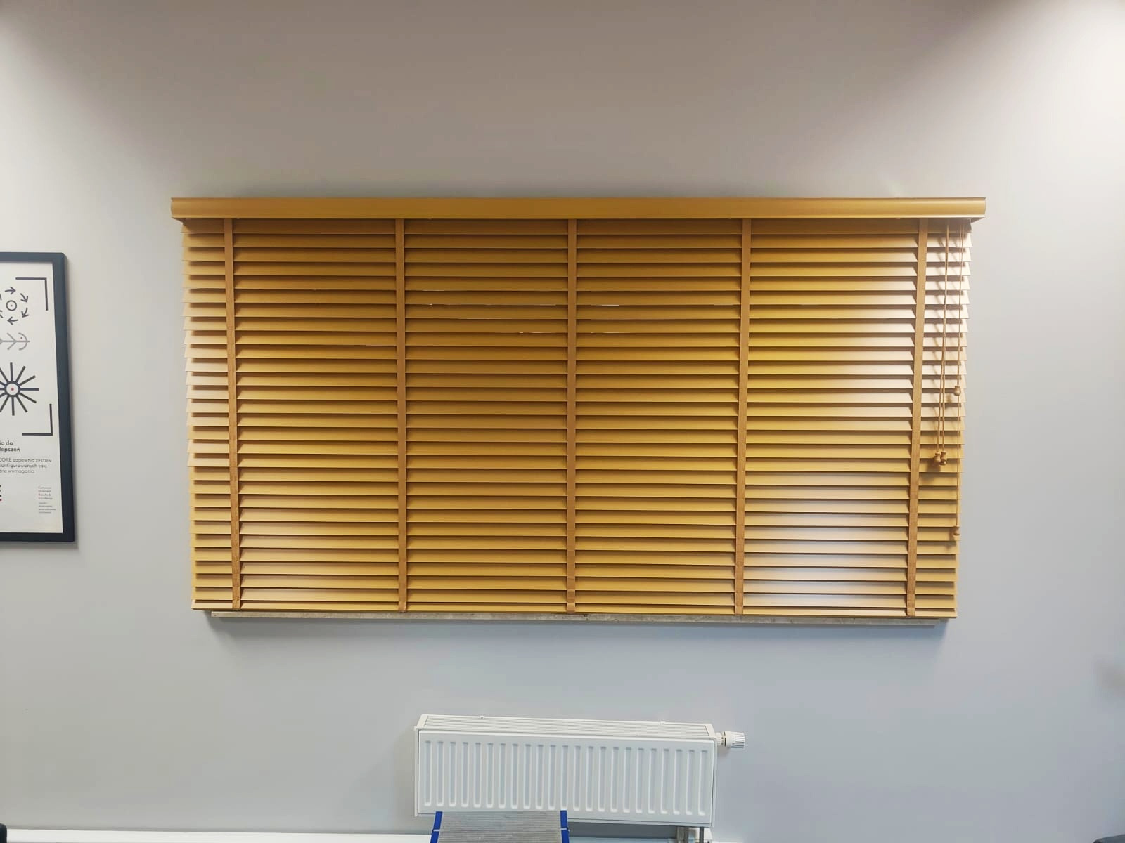 Zamknięte drewniane żaluzje w ciepłym odcieniu drewna, zamontowane w oknie na tle szarej ściany z grzejnikiem pod spodem i plakatem po lewej stronie.