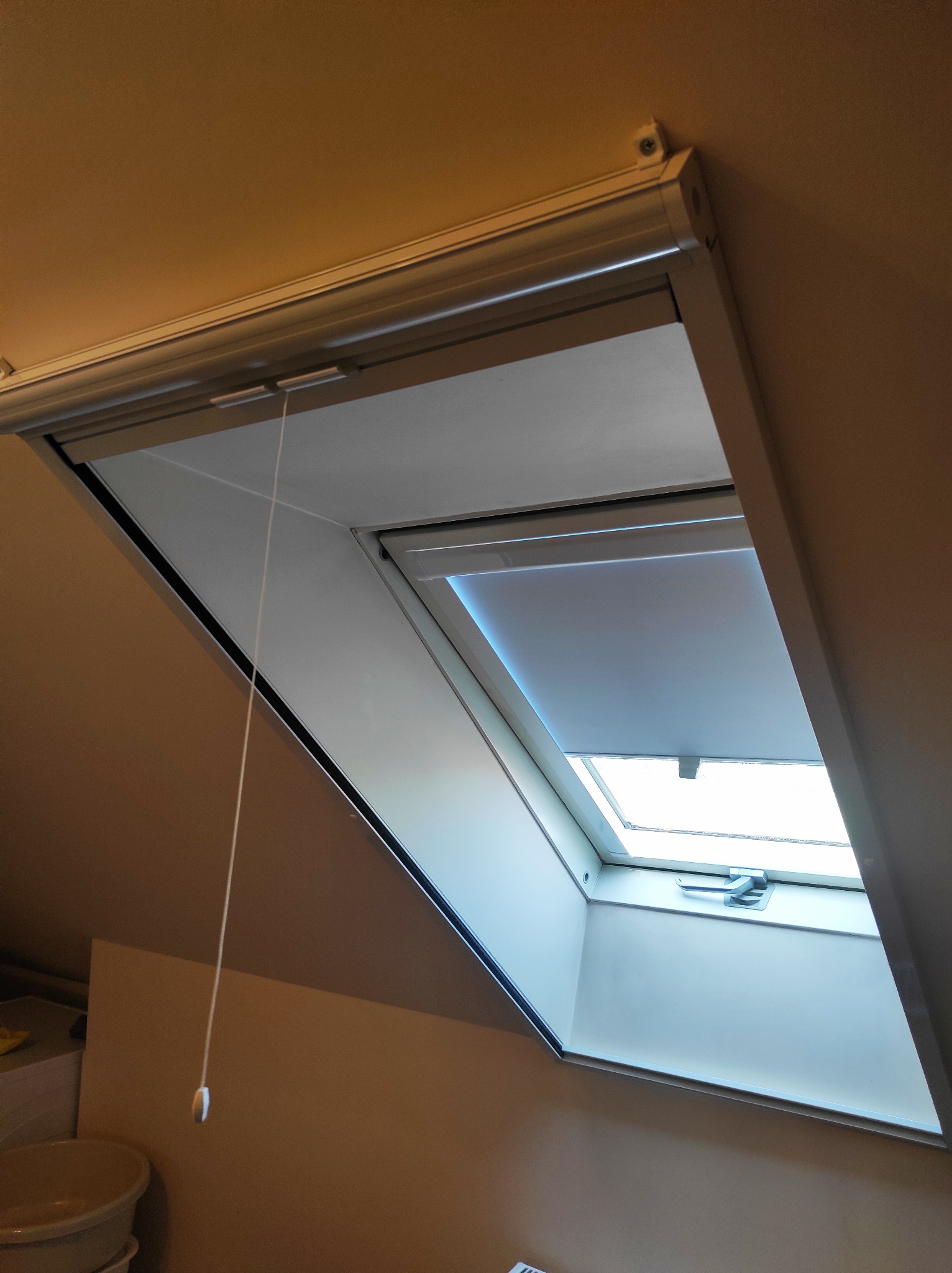 Szara roleta wewnętrzna zamontowana w oknie dachowym, częściowo zasłonięta, z widocznym mechanizmem sterującym sznurkiem.