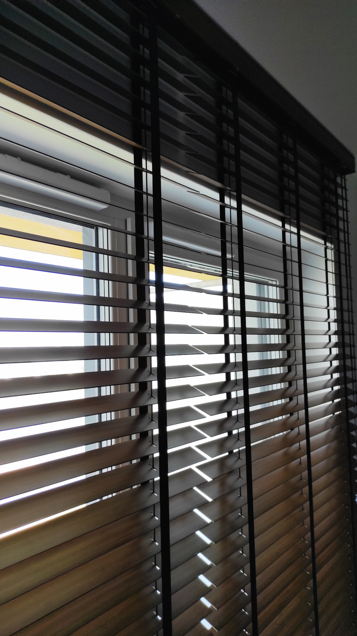 Częściowo zamknięte drewniane żaluzje z czarnymi sznurkami na oknie, wpadające promienie słoneczne tworzą wzory światła i cienia.