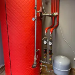 Czerwony, izolowany zbiornik akumulacyjny ciepła z podłączonymi rurami, zaworami i manometrem, obok szarego naczynia przeponowego w pomieszczeniu z panelami podłogowymi.