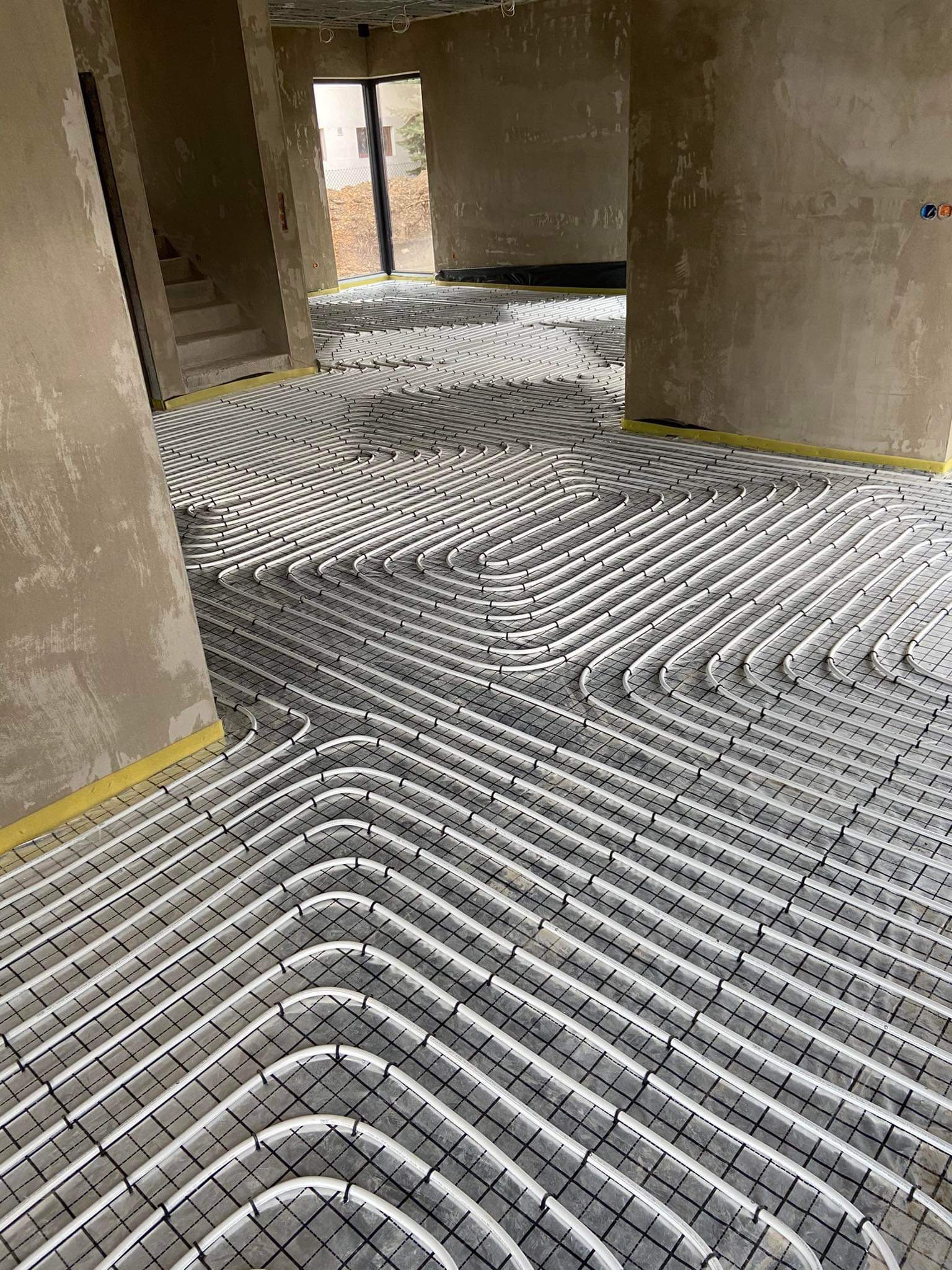 Rozłożone białe rury ogrzewania podłogowego na czarnej siatce w surowym wnętrzu budynku w trakcie budowy, widoczne fragmenty ścian i schodów.