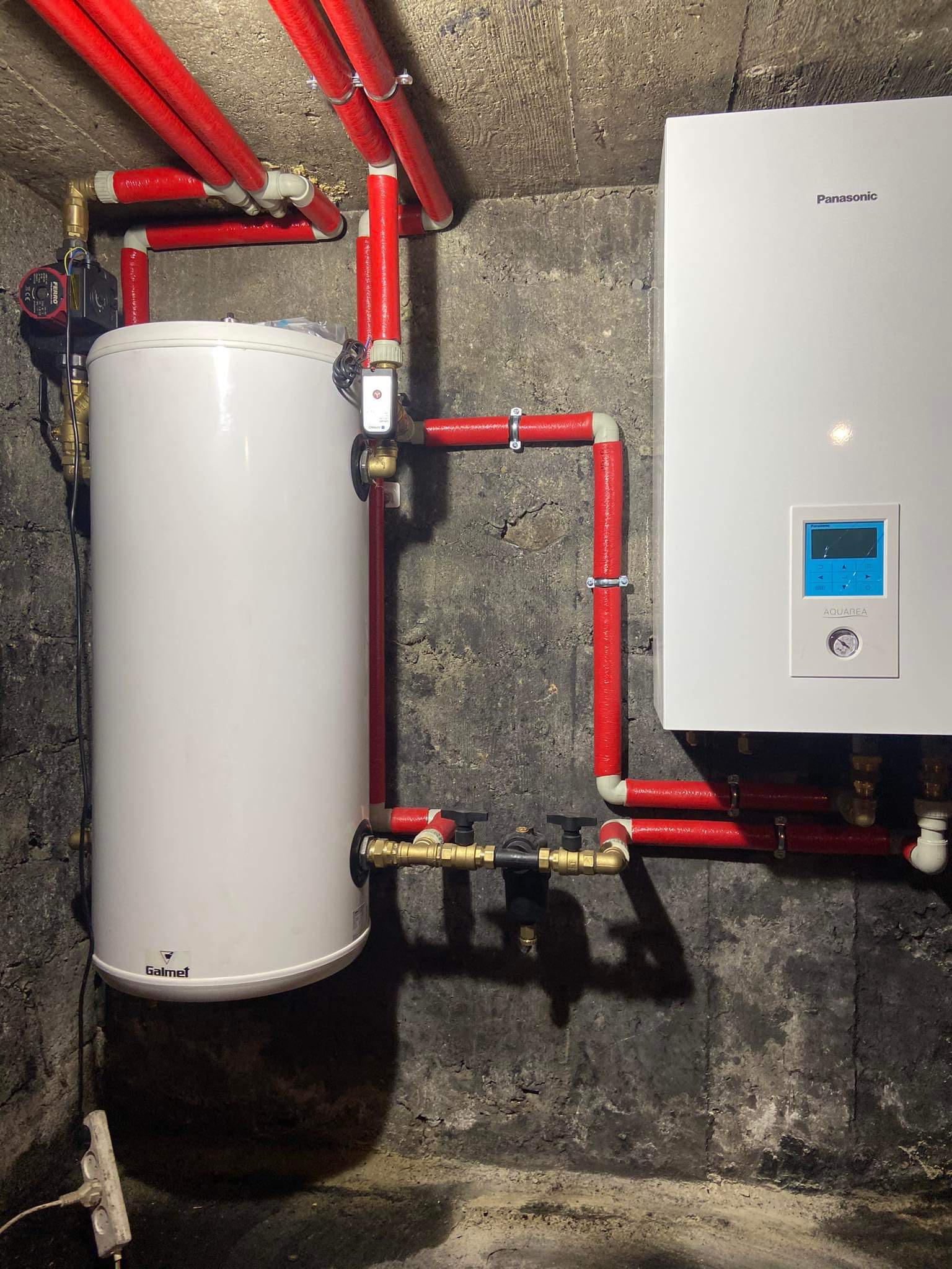 Bojler Galmet i piec gazowy Panasonic Aquarea połączone rurami w izolacji termicznej, zamontowane na tle surowej, betonowej ściany piwnicy.