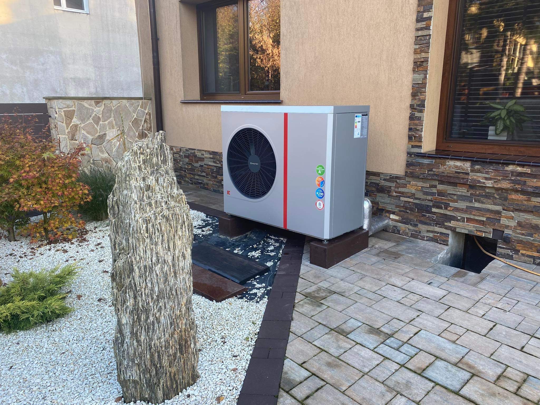 Zewnętrzna pompa ciepła powietrze-woda zamontowana na tarasie z kostki brukowej, obok dekoracyjny kamień w ogrodzie.