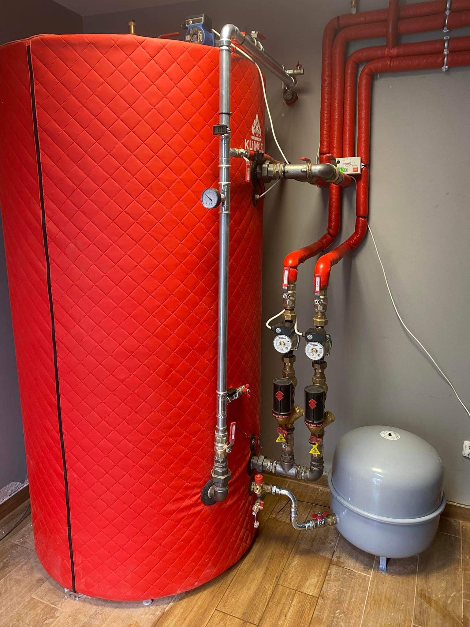 Czerwony, izolowany zbiornik akumulacyjny ciepła z podłączonymi rurami, zaworami i manometrem, obok szarego naczynia przeponowego w pomieszczeniu z panelami podłogowymi.