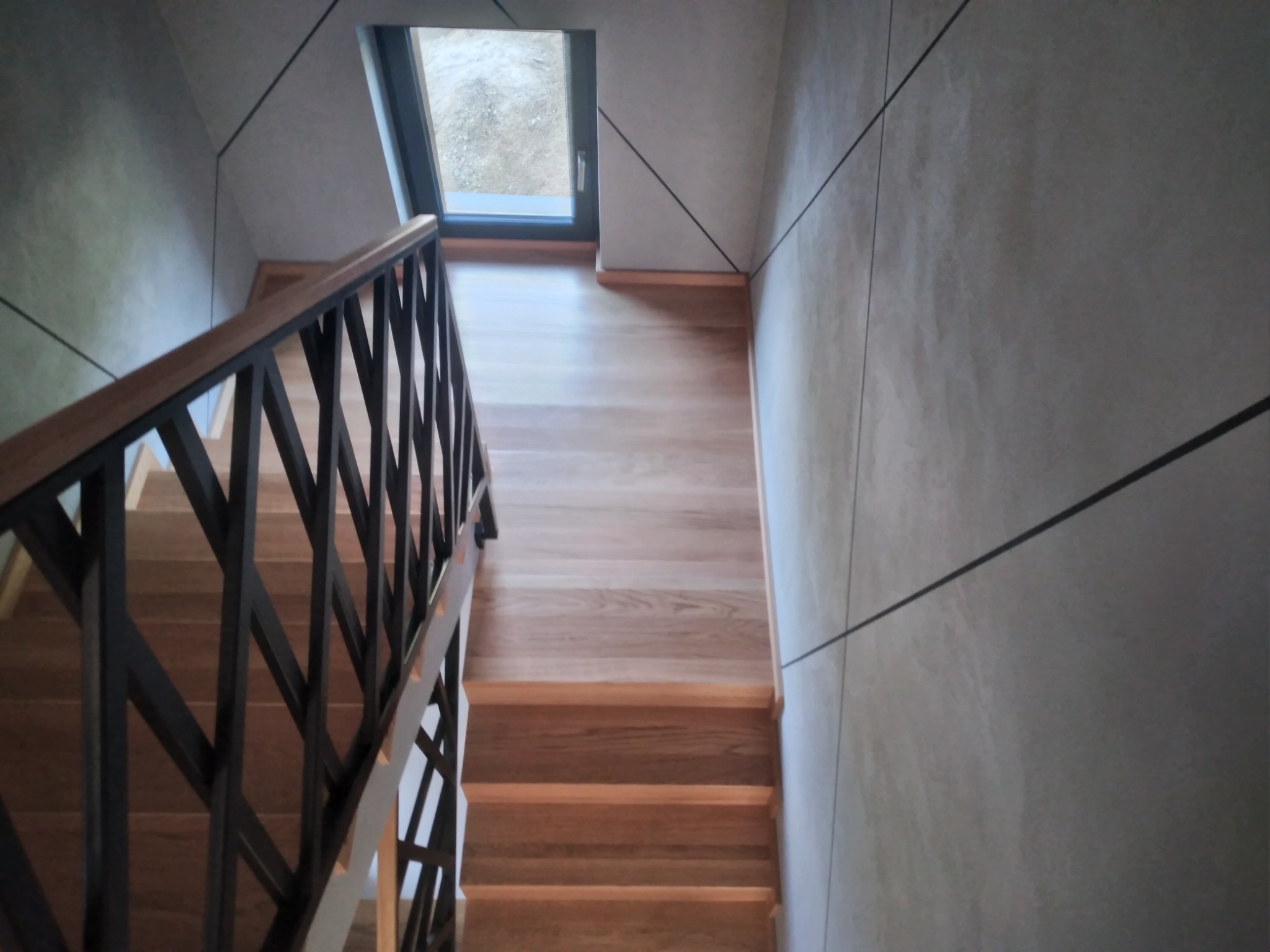 Widok z góry na drewniane stopnie z czarną, geometryczną balustradą w nowoczesnym wnętrzu z oknem.