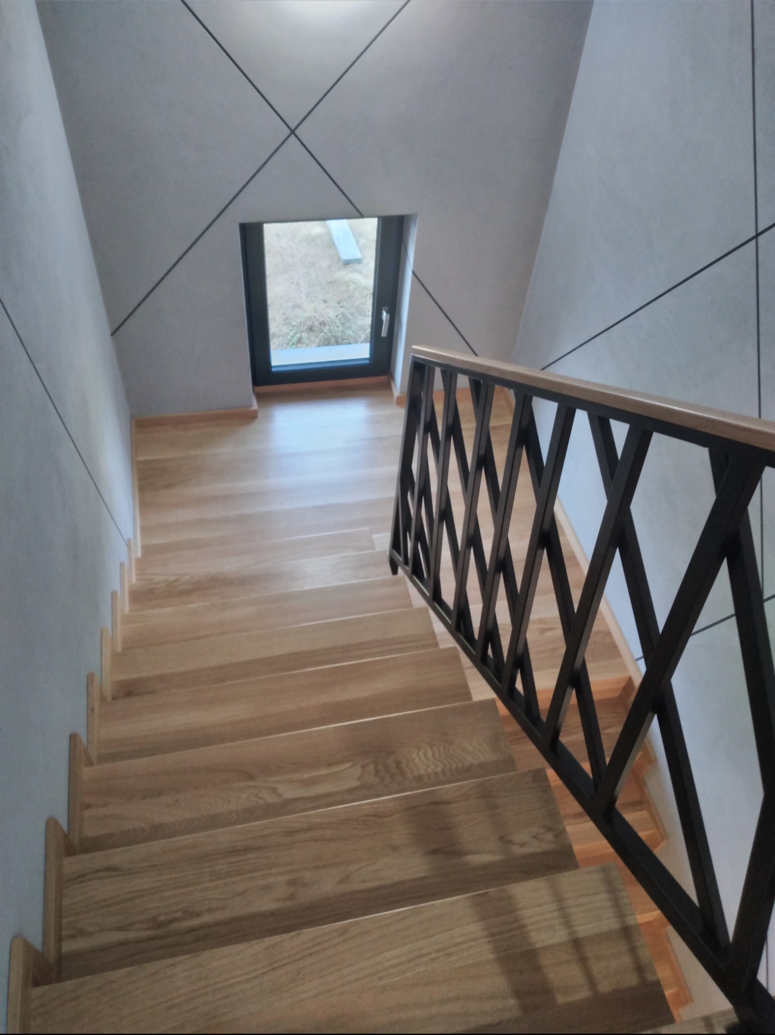 Wnętrze z drewnianymi schodami z metalową balustradą o geometrycznym wzorze, widok z góry na dół, ściany w jasnym odcieniu szarości z czarnymi liniami tworzącymi wzór, okno w tle.
