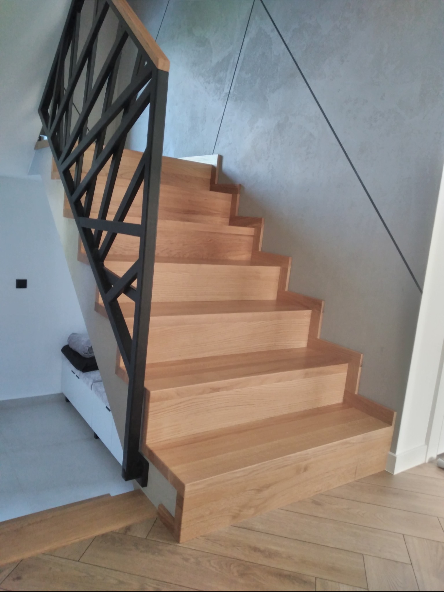 Jasne, drewniane stopnie z czarną, geometryczną balustradą, prowadzące do góry w nowoczesnym wnętrzu z szarą ścianą akcentową.