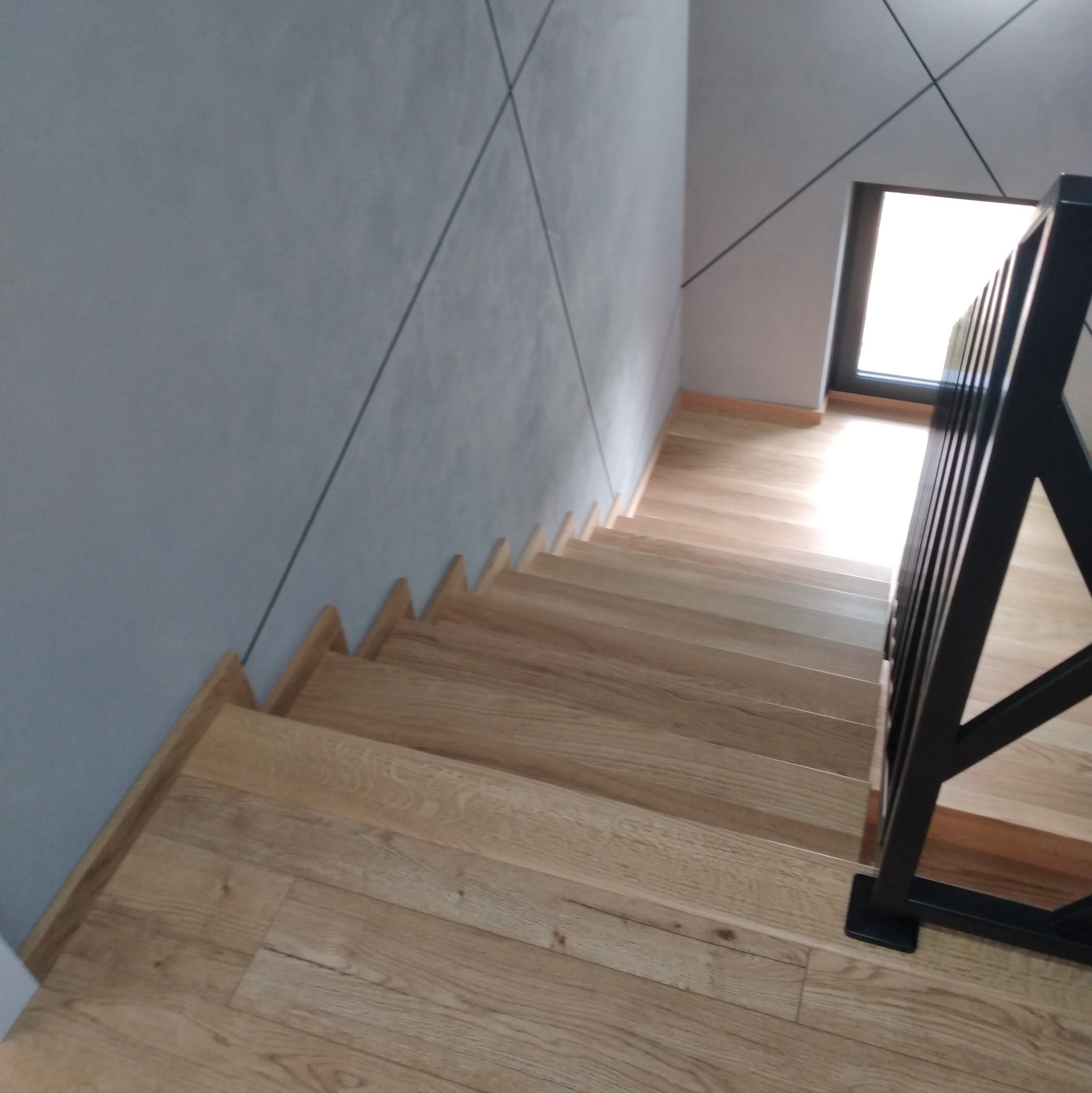 Widok z góry na drewniane schody z czarną, metalową balustradą, ściana w kolorze szarym z geometrycznym wzorem.